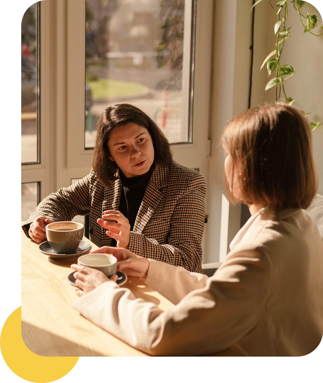 Two women talking over coffee by window.